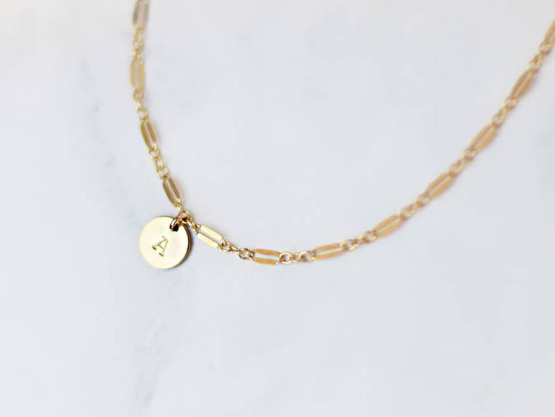 May include: A gold chain necklace with a small round pendant featuring the letter "A" engraved on it.