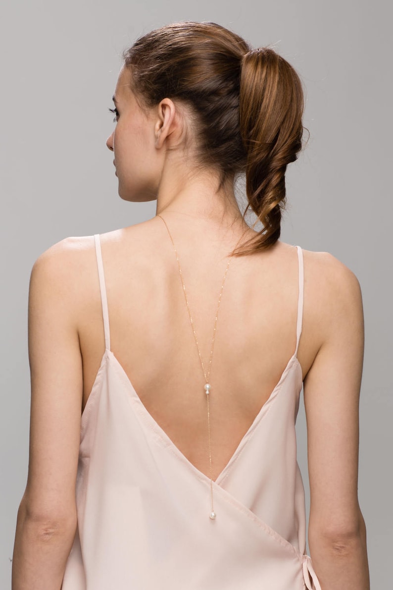 May include: A woman wearing a light pink, sleeveless top with a gold chain necklace with two pearl accents hanging down the back.