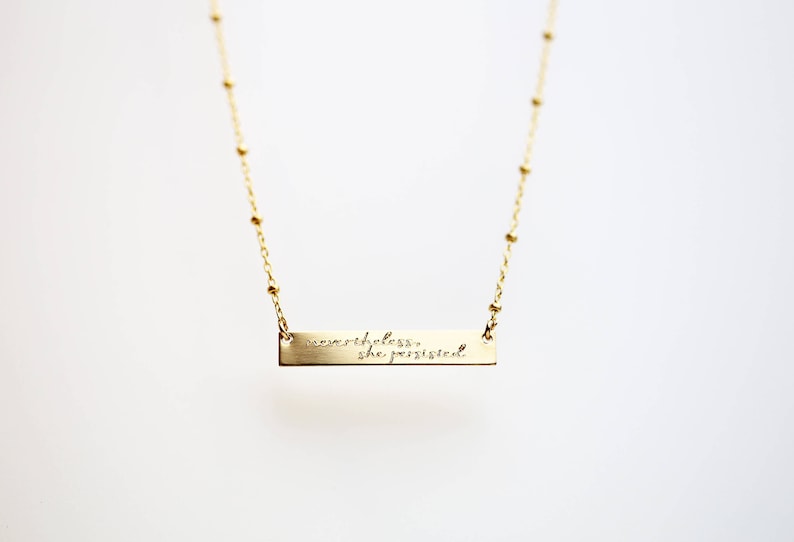 May include: A gold necklace with a bar pendant that reads "nevertheless, she persisted."