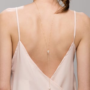 May include: A long gold chain necklace with two white pearls hanging from it. The necklace is worn on the back of a person's neck.