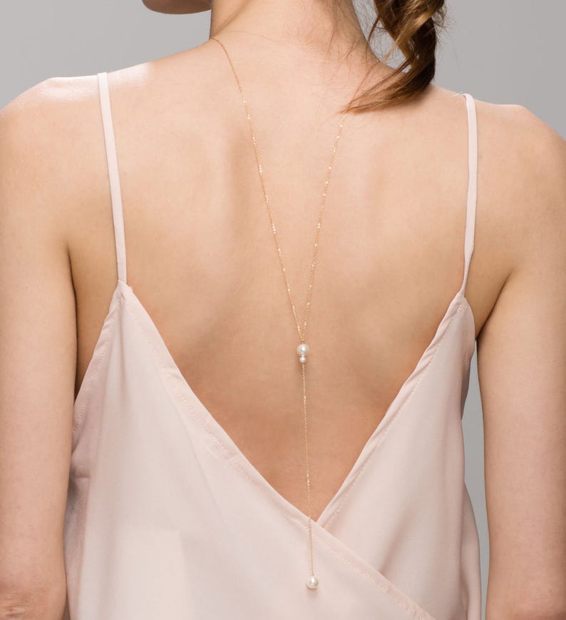 May include: A long gold chain necklace with two white pearls hanging from it. The necklace is worn on the back of a person's neck.