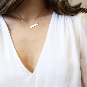 May include: A gold bar necklace with a delicate chain. The necklace is worn by a person with long brown hair.