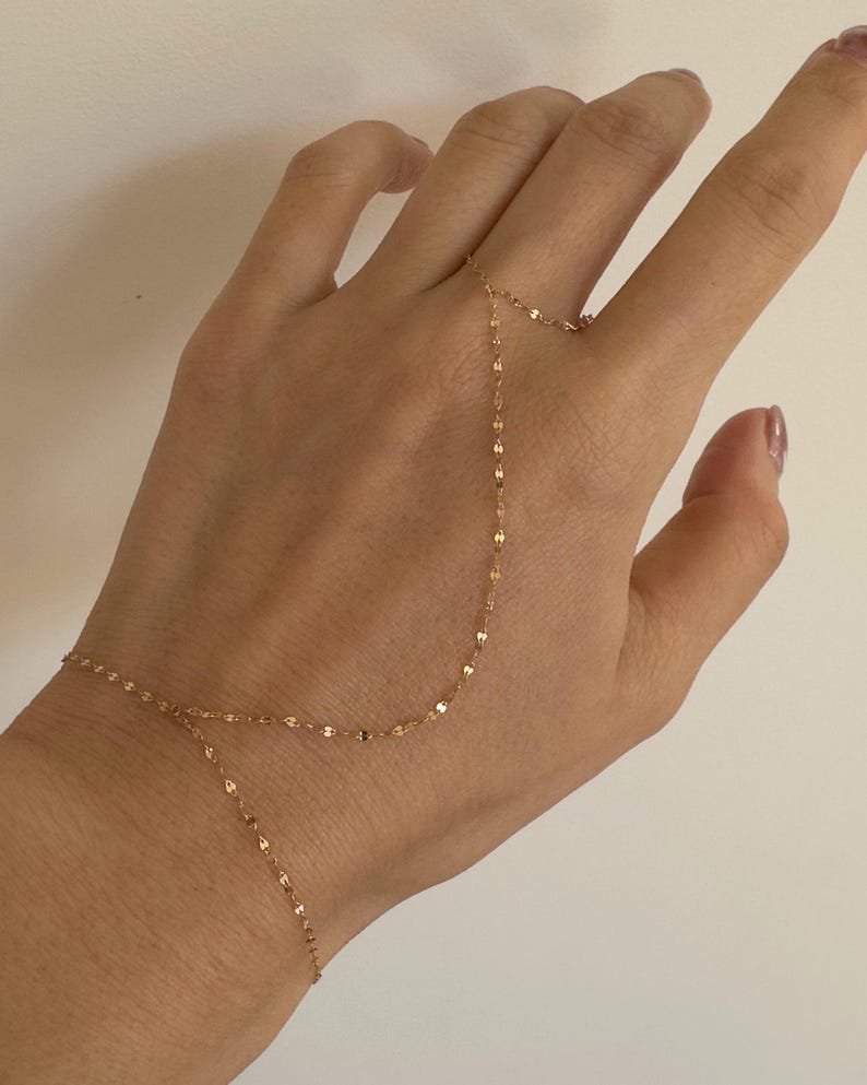 K&ouml;nnte beinhalten: Eine zarte goldene Handkette, die eine feine Kette aufweist, die ein Armband mit einem Ring verbindet. Die Kette besteht aus kleinen, flachen, reflektierenden Gliedern. Der Schmuck wird vor einem neutralen Hintergrund pr&auml;sentiert.