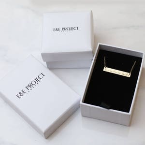 May include: A gold bar necklace with the text "If lost, please return to" on a black velvet background inside a white box with the text "E&E Project Jewelry".