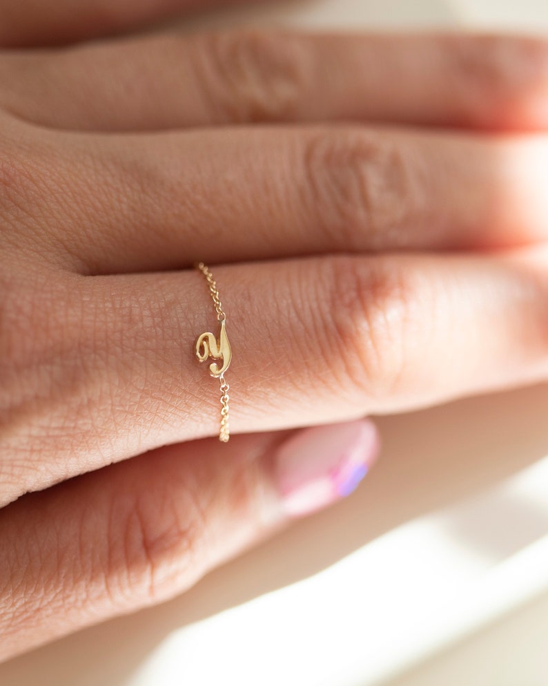 Puede incluir: Un delicado anillo de cadena de oro con un peque&ntilde;o dije con la letra 'y'. El anillo se lleva en un dedo con esmalte de u&ntilde;as rosa claro y azul.
