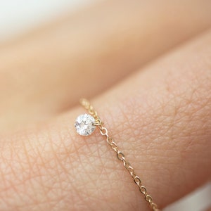 May include: A delicate gold chain ring with a single, sparkling diamond pendant. The ring is simple and elegant, featuring a small, round diamond suspended from the chain. The ring is shown on a person's finger.