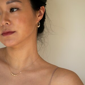 May include: A gold necklace with a delicate chain and a row of small, round beads. The necklace is worn by a person with dark hair and a light complexion.