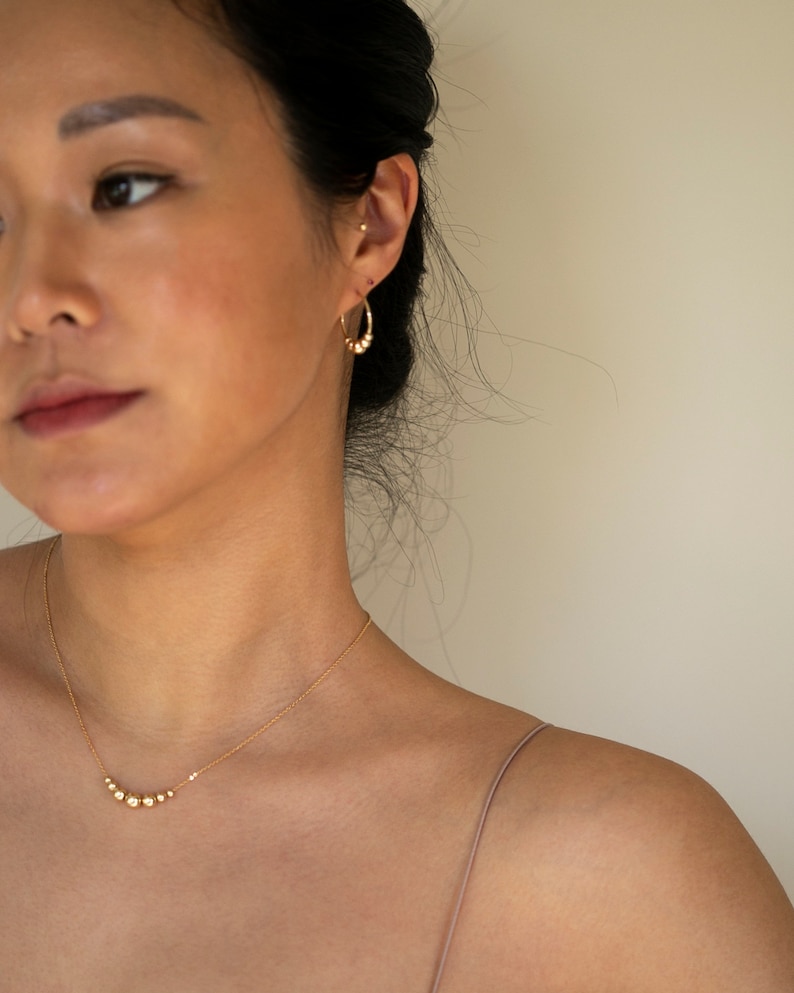 May include: A gold necklace with a delicate chain and a row of small, round beads. The necklace is worn by a person with dark hair and a light complexion.