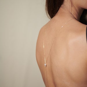 May include: A gold chain necklace with two pearl accents hangs down the back of a person's neck. The necklace is worn with a black top.