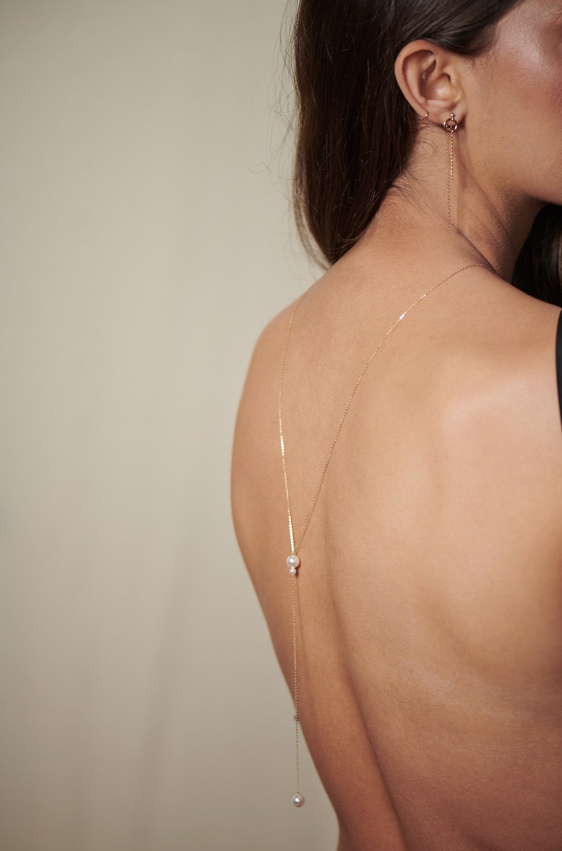 May include: A gold chain necklace with two pearl accents hangs down the back of a person's neck. The necklace is worn with a black top.