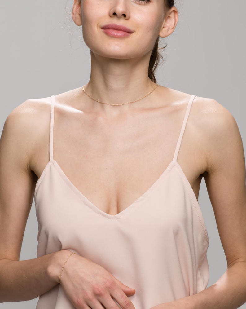 May include: A woman wearing a light pink sleeveless top with a delicate gold chain necklace. The necklace is thin and has a simple design.