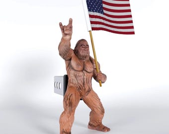 May include: A brown, furry Bigfoot figurine attached to a silver hitch cover. The figure stands in a wide stance, holding a gold pole with an American flag in one hand and making a rock-and-roll hand gesture with the other raised hand.