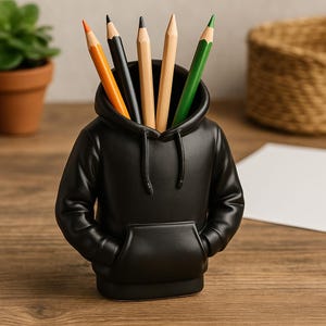 Può includere: Porta matite nero a forma di felpa con cappuccio con cinque matite colorate che sporgono dalla parte superiore. Le matite sono arancioni, nere, marroni, marroni e verdi.