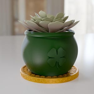 May include: A green pot with a four-leaf clover design, holding a light green succulent plant. The pot sits on a gold-colored, circular coaster with a repeating pattern. The plant pot is approximately 4 inches in diameter.