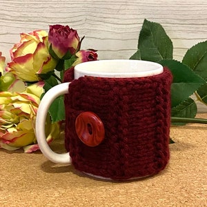 May include: A white ceramic mug with a burgundy knitted cozy and a large, round button. The cozy is wrapped around the mug and has a textured appearance. The mug is placed in front of a floral arrangement with yellow and red roses.