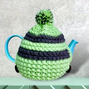 May include: A light blue teapot with a green and navy blue crocheted tea cozy. The cozy has horizontal stripes and a green pom-pom on top. The teapot is on a light green surface.