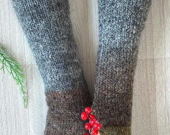May include: A pair of knitted arm warmers in shades of gray, brown, and olive green. The arm warmers are made of a thick, textured yarn and have a ribbed cuff. A sprig of red berries is held between the fingers.
