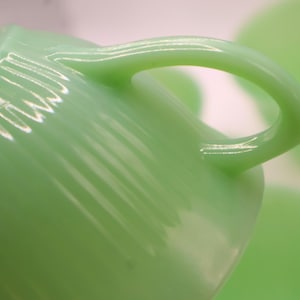 May include: Close-up of a light green teacup with a curved handle. The cup has a smooth, glossy finish and a slightly ribbed texture. The handle is rounded and appears sturdy. The background is blurred, suggesting a shallow depth of field.
