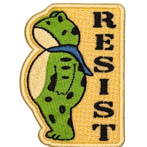 May include: Embroidered patch featuring a green frog with black spots and a blue scarf. The frog is positioned next to a yellow rectangle with the word "RESIST" in black capital letters. The patch has a gold border.