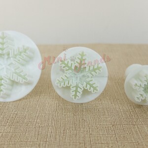 May include: Three white plastic snowflake cookie cutters of different sizes. The largest cutter is 5 cm in diameter, the middle cutter is 3.8 cm in diameter, and the smallest cutter is 2.5 cm in diameter.