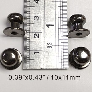 Könnte beinhalten: Vier schwarze Metallknöpfe mit runder Oberseite und flachem Boden. Die Knöpfe sind 0,39" x 0,43" / 10 x 11 mm groß.