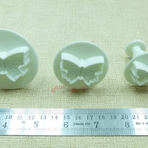 May include: Three white plastic butterfly-shaped cookie cutters of different sizes. The largest cutter is 5 inches wide, the middle cutter is 3 inches wide, and the smallest cutter is 2 inches wide.