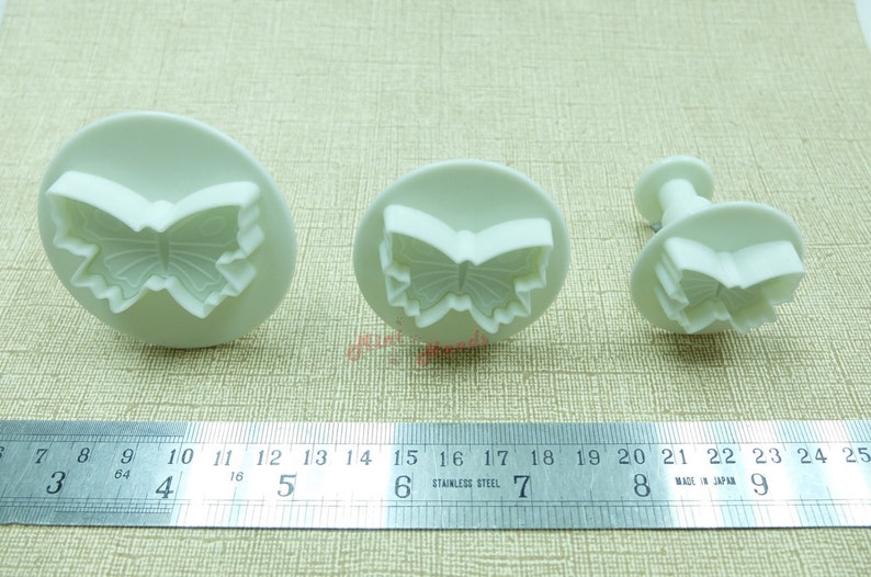 May include: Three white plastic butterfly-shaped cookie cutters of different sizes. The largest cutter is 5 inches wide, the middle cutter is 3 inches wide, and the smallest cutter is 2 inches wide.