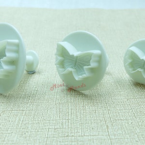 May include: Three white plastic butterfly cookie cutters of different sizes. The cutters have a raised butterfly design with a scalloped edge.