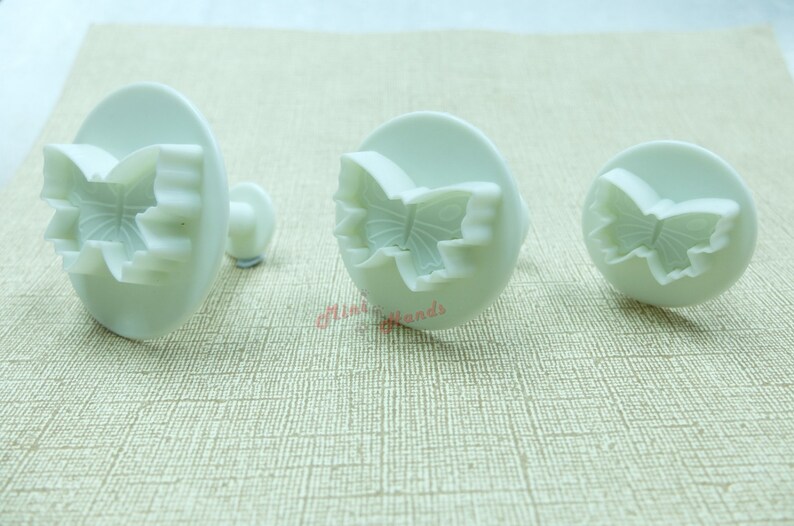 May include: Three white plastic butterfly cookie cutters of different sizes. The cutters have a raised butterfly design with a scalloped edge.