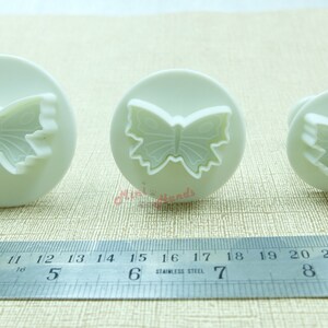 May include: Three white plastic butterfly cookie cutters of different sizes. The cutters are on a white surface with a ruler for scale.