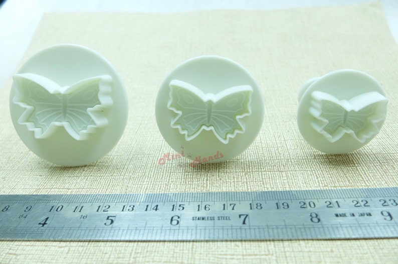May include: Three white plastic butterfly cookie cutters of different sizes. The cutters are on a white surface with a ruler for scale.