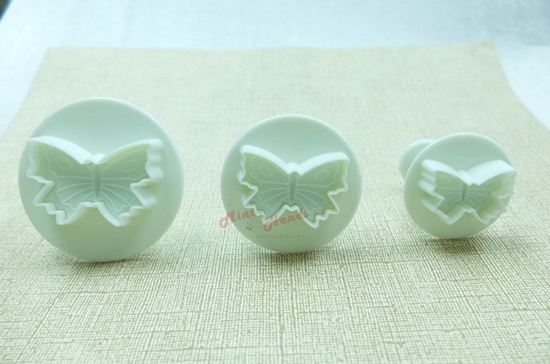 May include: Three white plastic butterfly-shaped cookie cutters of different sizes. The cutters are on a beige surface.