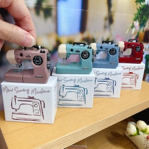 Könnte beinhalten: Vier Miniatur-Nähmaschinen in verschiedenen Farben, darunter rosa, blau, hellblau und rot, werden auf einem Holzregal ausgestellt. Jede Maschine befindet sich auf einem weißen Kasten mit dem aufgedruckten Text "Mini Sewing Machine". Die Kisten haben eine Grafik einer Nähmaschine darauf.