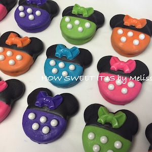 May include: Assortment of nine decorated chocolate-covered Oreos shaped like Mickey Mouse heads with bows. The Oreos are decorated in various colors, including purple, green, orange, pink, and blue. Each Oreo has white chocolate dots on the front.