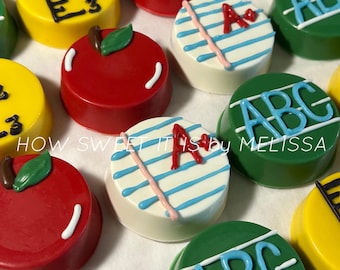 Puede incluir: Primer plano de varios dulces decorados con temática escolar. Algunos tienen forma de manzana roja, otros son amarillos con diseños de regla, y algunos son blancos con rayas azules y una "A" roja. Los dulces verdes tienen "ABC" en azul. El texto dice "HOW SWEET IT IS by MELISSA".