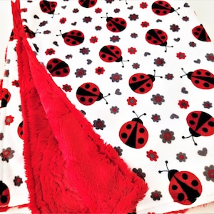 May include: A white blanket with a red, black, and gray ladybug and flower pattern. The blanket has a soft, red, plush backing. The ladybugs have black bodies with red spots. The flowers are red and gray.