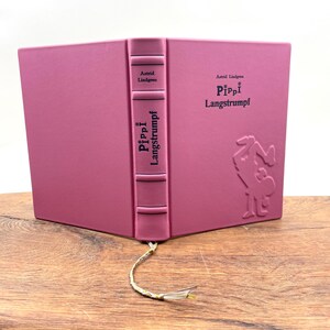Puede incluir: Un libro de tapa dura rosa con el t&iacute;tulo "Pippi Langstrumpf" en relieve en la portada y el lomo, junto con el nombre del autor, Astrid Lindgren. El libro tiene un marcap&aacute;ginas con borla dorada. La portada tiene una ilustraci&oacute;n en relieve.