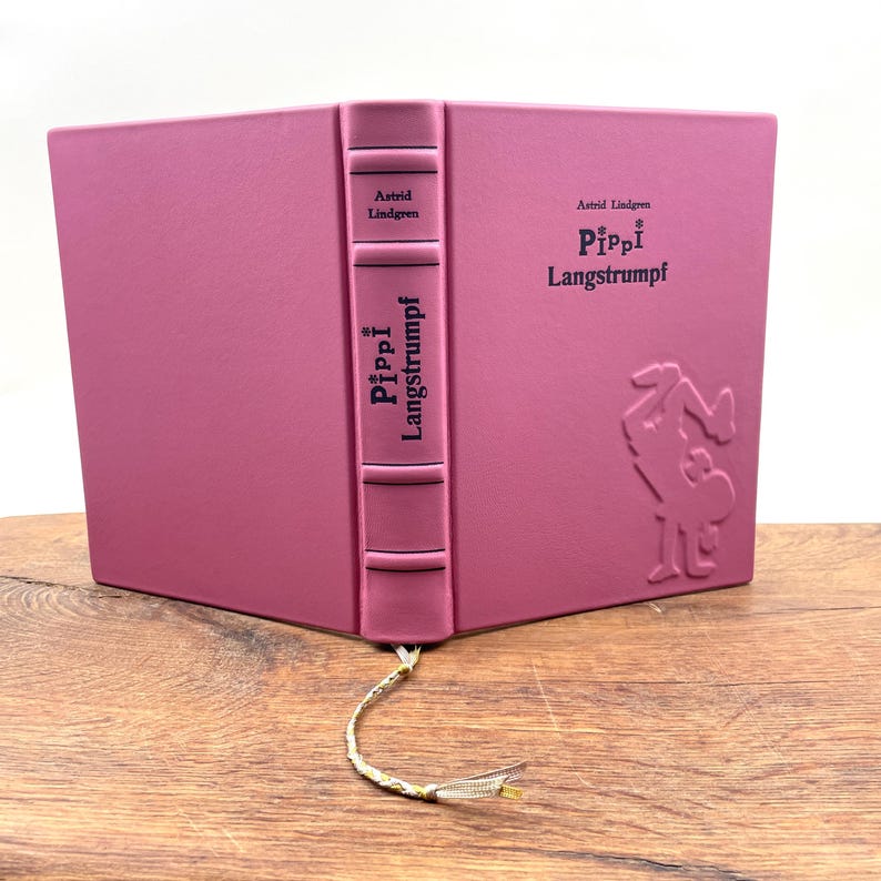 Puede incluir: Un libro de tapa dura rosa con el t&iacute;tulo "Pippi Langstrumpf" en relieve en la portada y el lomo, junto con el nombre del autor, Astrid Lindgren. El libro tiene un marcap&aacute;ginas con borla dorada. La portada tiene una ilustraci&oacute;n en relieve.