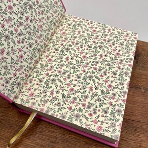 Puede incluir: Diario abierto con una cubierta rosa y p&aacute;ginas con estampado floral. Las p&aacute;ginas presentan un fondo crema con un delicado patr&oacute;n de flores y hojas rosas, grises y verdes. Se ve una cinta dorada como marcap&aacute;ginas.
