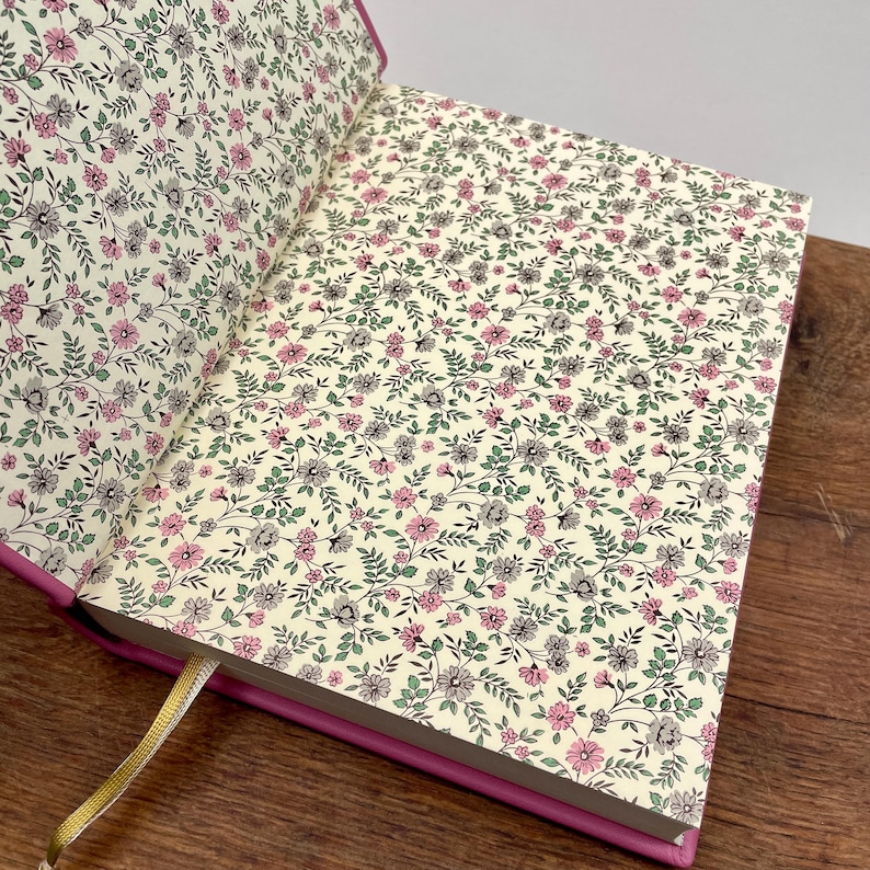 Puede incluir: Diario abierto con una cubierta rosa y p&aacute;ginas con estampado floral. Las p&aacute;ginas presentan un fondo crema con un delicado patr&oacute;n de flores y hojas rosas, grises y verdes. Se ve una cinta dorada como marcap&aacute;ginas.