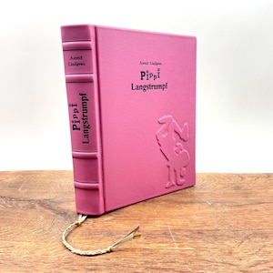 Puede incluir: Un libro rosa de tapa dura titulado "Pippi Langstrumpf" de Astrid Lindgren. El libro tiene ilustraciones y texto en relieve en la portada y el lomo. Se adjunta un marcap&aacute;ginas decorativo. El libro se muestra sobre una superficie de madera.
