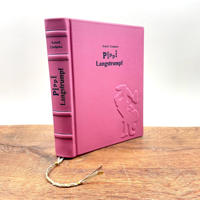Puede incluir: Un libro rosa de tapa dura titulado "Pippi Langstrumpf" de Astrid Lindgren. El libro tiene ilustraciones y texto en relieve en la portada y el lomo. Se adjunta un marcap&aacute;ginas decorativo. El libro se muestra sobre una superficie de madera.