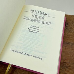Puede incluir: Un libro abierto con el t&iacute;tulo "Pippi Langstrumpf" de Astrid Lindgren. Las p&aacute;ginas son de color crema y la portada es rosa vibrante. El texto est&aacute; en alem&aacute;n, con el nombre del editor en la parte inferior.