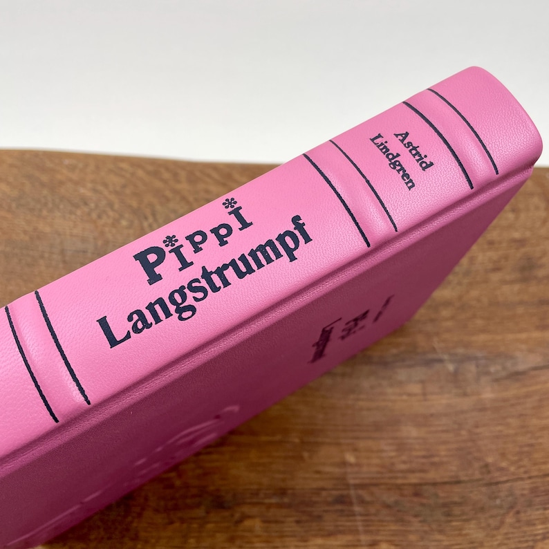 Puede incluir: Un libro de tapa dura rosa titulado "Pippi Langstrumpf" de Astrid Lindgren. El lomo del libro presenta el t&iacute;tulo en letras azul oscuro y el nombre del autor. El libro est&aacute; sobre una superficie de madera.