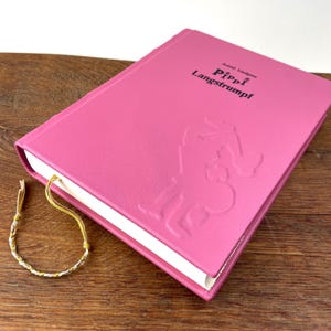 Puede incluir: Un libro rosa de tapa dura titulado "Pippi Langstrumpf" con arte de personajes en relieve. El libro tiene una cinta dorada como marcap&aacute;ginas y descansa sobre una superficie de madera. Una peque&ntilde;a pulsera trenzada con hilos dorados y plateados est&aacute; al lado del libro.