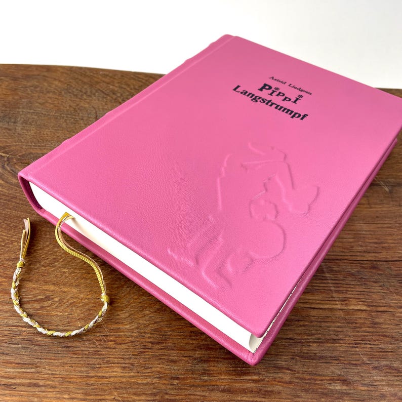 Puede incluir: Un libro rosa de tapa dura titulado "Pippi Langstrumpf" con arte de personajes en relieve. El libro tiene una cinta dorada como marcap&aacute;ginas y descansa sobre una superficie de madera. Una peque&ntilde;a pulsera trenzada con hilos dorados y plateados est&aacute; al lado del libro.