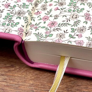 Puede incluir: Primer plano de un diario con una cubierta rosa y p&aacute;ginas interiores con estampado floral. Las p&aacute;ginas presentan un dise&ntilde;o de flores rosas, grises y blancas con hojas verdes. Se ve un marcap&aacute;ginas de cinta dorada y plateada.