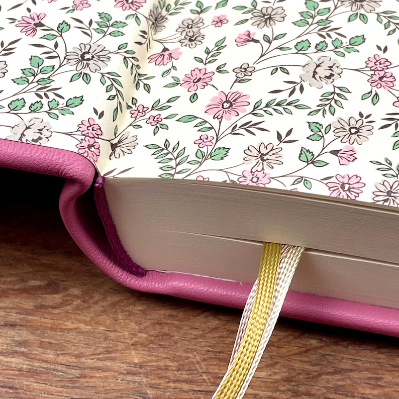 Puede incluir: Primer plano de un diario con una cubierta rosa y p&aacute;ginas interiores con estampado floral. Las p&aacute;ginas presentan un dise&ntilde;o de flores rosas, grises y blancas con hojas verdes. Se ve un marcap&aacute;ginas de cinta dorada y plateada.