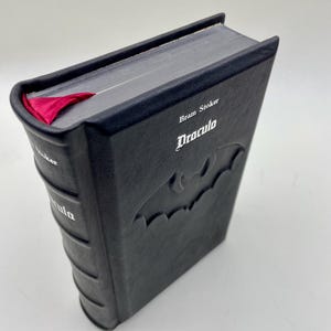 Könnte beinhalten: Ein schwarzes Hardcover-Buch mit dem Titel "Dracula" von Bram Stoker. Das Buch zeigt ein Fledermaus-Design und ein rotes Schleifen-Lesezeichen. Der Titel ist auf dem Buchrücken wiederholt. Das Buch ist vor einem weißen Hintergrund platziert.