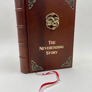 Könnte beinhalten: Ein braunes, ledergebundenes Buch mit dem Titel "The Neverending Story" mit goldenen Akzenten und dekorativem Eckdesign. Das Buch hat ein goldenes Emblem aus verschlungenen Schlangen und ein rot-weißes Leseband.